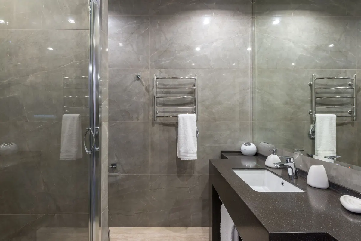 Modern bathroom with gray tiled walls, a walk-in glass shower, dark stone counter and folded white towels