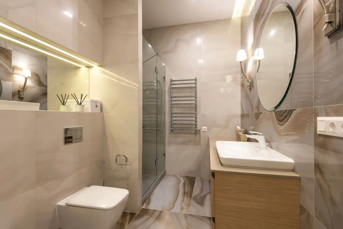 Marble-tiled bathroom with a round backlit mirror, wall-hung sink, glass shower enclosure and towel rail