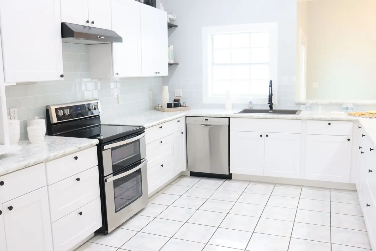 White kitchen with light tile flooring, stainless steel range, dishwasher and a window above the sink