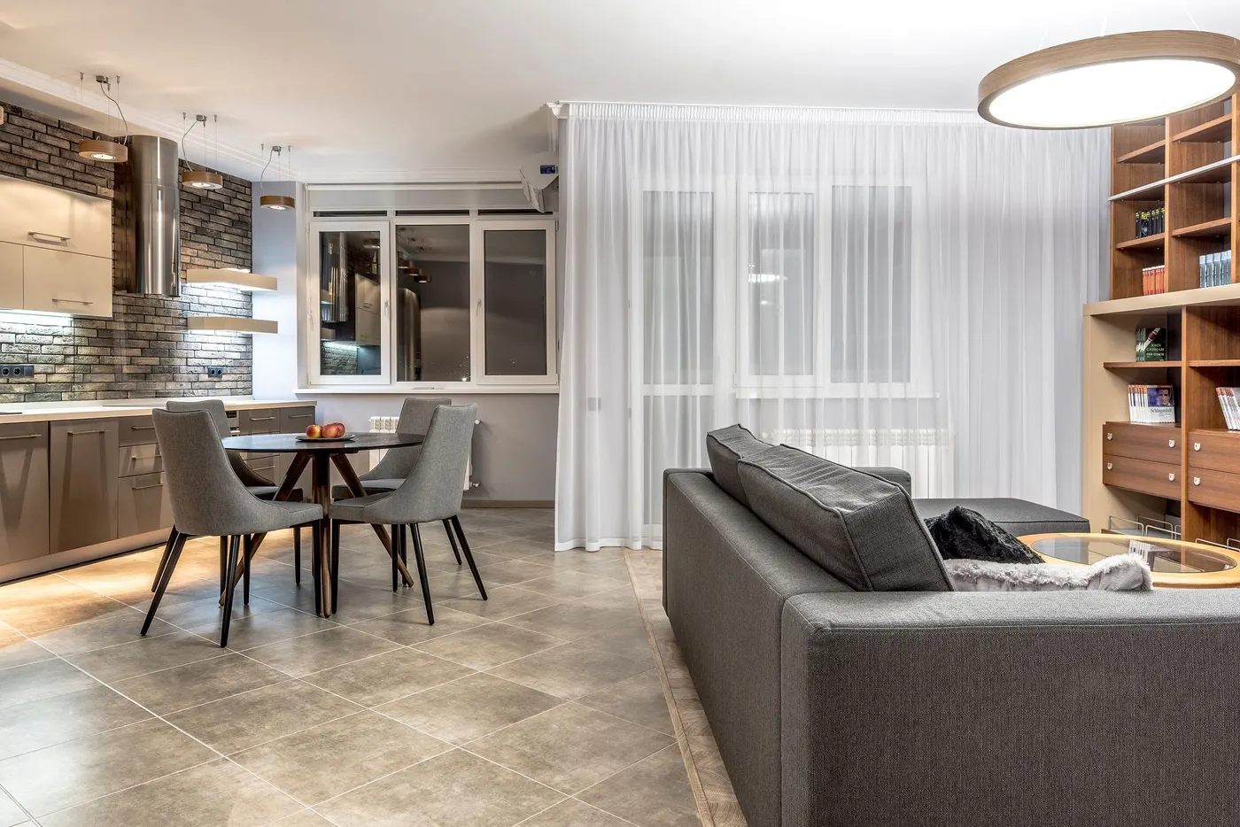 Open-plan living area with a gray sectional sofa, dining table, built-in shelving and a kitchen along the far wall